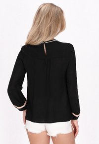 Blusa nera a maniche lunghe con tessuto texturizzato, dettaglio a ochetta nella parte posteriore e bordi colorati ai polsini. Abbinata a pantaloni shorts bianchi sfrangiati.