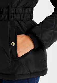 Mustikka musta takki, jossa on vyöllinen vyötärö, elastiset hihansuut ja sivutaskut hopeisilla painonapeilla. Sileä, kiiltävä kangas.