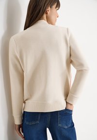 Femme aux longs cheveux bruns portant un pull côtelé couleur crème et un jean bleu foncé, debout avec une main dans la poche arrière.