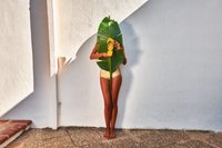 Costume da bagno giallo con ritagli laterali, tenuto con una grande foglia verde davanti, in piedi su una superficie di pietra testurizzata contro un muro bianco.