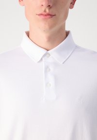Hvit polo skjorte med klassisk krage, tre knapper og en subtil brodert logo på brystet. Glatt bomullsstoff.