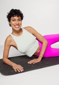 Camisola sem mangas cinza claro com nervuras, combinada com leggings rosas brilhantes de cintura alta, deitadas sobre um tapete de fitness preto texturizado com manchas coloridas.