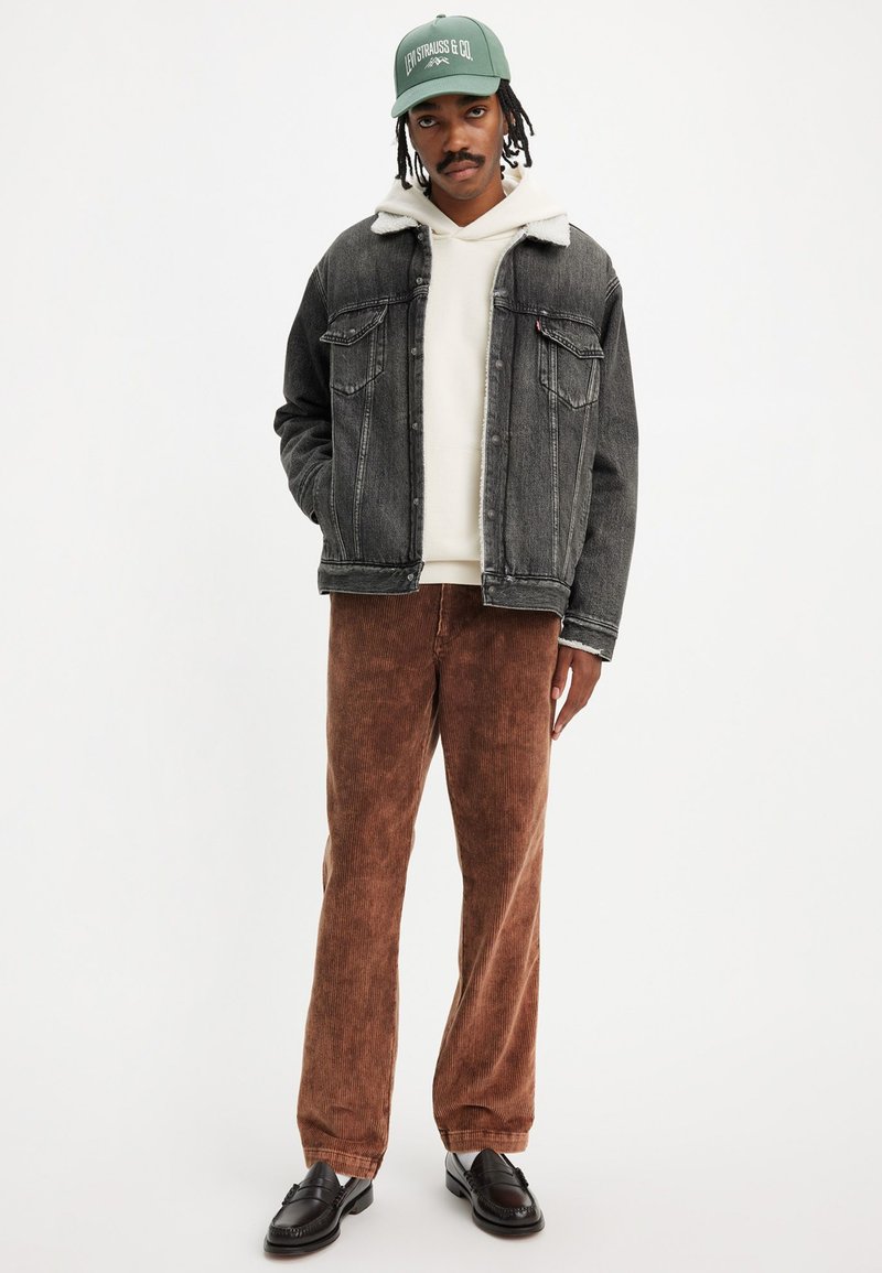 Mann trägt eine grüne Levi’s-Cap, eine schwarze Jeansjacke mit weißem Fleece-Kragen, einen cremefarbenen Hoodie, braune Cordhosen und schwarze Slipper vor weißem Hintergrund.
