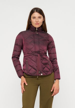 Femme portant une veste matelassée violette avec un col montant et une ceinture, associée à un pantalon vert olive, debout devant un fond uni.