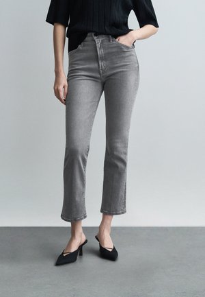 Žena oblečená v sivých skrátených džínsoch, čiernom tričku s krátkym rukávom a čiernych lodičkách s otvorenou pätou a špicatou špičkou stojaca na sivom podlaží.