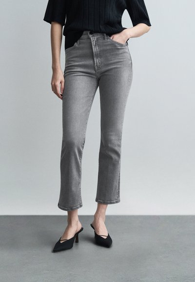 Femme portant un jean gris raccourci, un haut noir à manches courtes et des escarpins slingback noirs à bouts pointus, debout sur un sol gris.