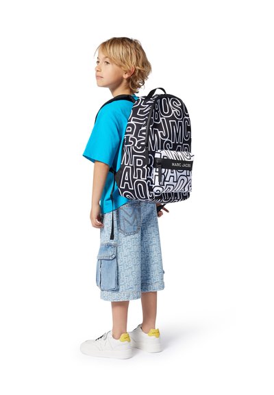 Sac à dos noir et blanc avec lettrage en gras, sangles réglables et une poche avant. Porté par un enfant en chemise bleue et shorts en denim.