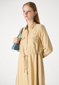 Beżowa sukienka z długim rękawem, z guzikami z przodu, dwoma kieszeniami na piersi i ściąganym paskiem w talii. Modelka trzyma niebieską, trójkątną torbę.