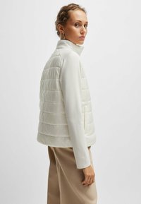 Giacca a vento bianca con una texture liscia e trapuntata, colletto alto e maniche lunghe, abbinata a pantaloni beige. Design minimalista.