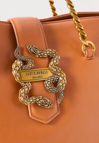 Tan leather tote bag featuring a prominent gold snake-shaped hardware detail with an engraved plate reading "JUST CAVALLI MILANO."