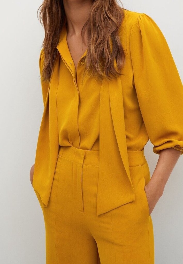 Femme portant un chemisier jaune moutarde à manches bouffantes et un pantalon taille haute assorti, les mains dans les poches, debout devant un fond uni.