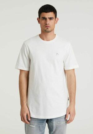 T-shirt bianco di cotone con maniche corte, scollatura rotonda e un piccolo logo geometrico ricamato sul lato sinistro del petto. Indossato con jeans chiari strappati.