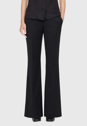 Woman wearing black high-waist, wide-leg trousers paired with black shoes and a black top, standing with one hand in pocket.