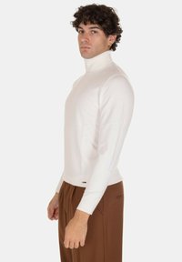 Maglione bianco a collo alto realizzato in morbido tessuto a maglia, con design aderente e dettagli a coste, abbinato a pantaloni marroni.