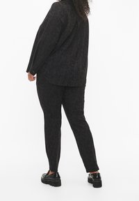 Person med lockigt hår som bär en svart mönstrad långärmad skjorta och matchande byxor, står med ryggen mot kameran, svarta platåskor synliga.