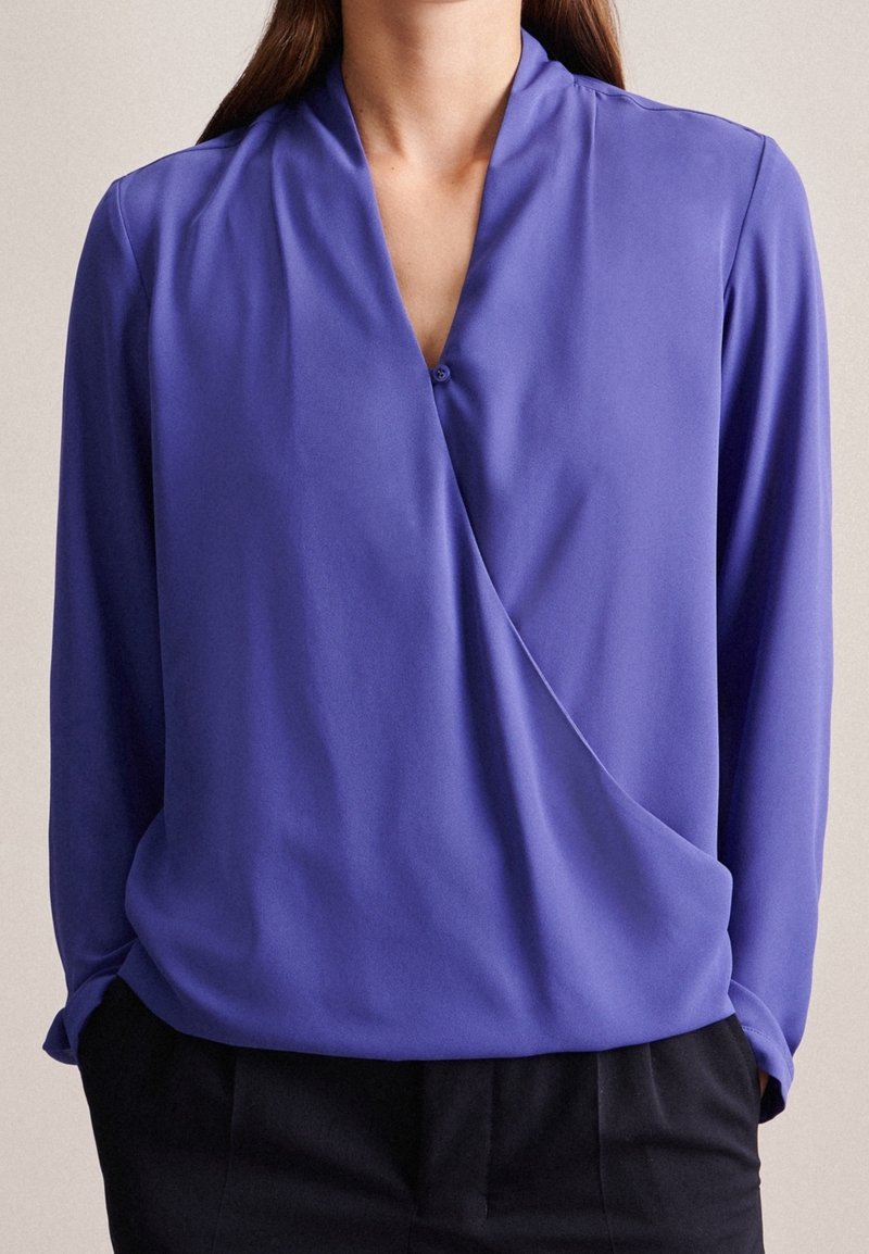 Femme portant un chemisier violet à manches longues style portefeuille, rentré dans un pantalon noir, debout les mains dans les poches.