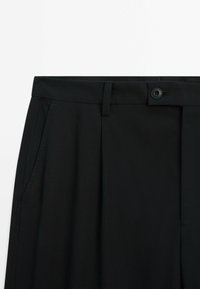 Black tailored pants with a smooth texture, featuring a button closure, front pleats, and side pockets.