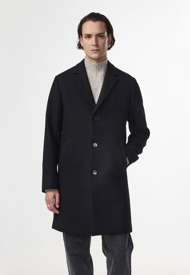 Musta villakangas takki, jossa on nuolimallinen kaulus, viisi nappia ja sivutaskut. Yllä vaaleanharmaa pooloneule ja harmaat farkut.