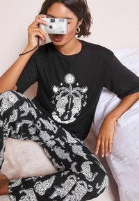 Tricou negru cu grafică albă ce ilustrează lei de mare, o palmă și un soare. Pantaloni negri cu imprimeu alb de leopard. Cameră argintie ținută în față.
