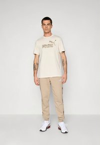 Beige bomulls-t-shirt med "PUMA"-logotyp i en mörkare tryck, kombinerad med beige sweatpants med grafiska detaljer på sidan och vita sneakers.