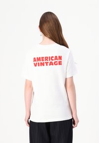 Balta kokvilnas T-kreklu ar īsām piedurknēm un apaļu kakla izgriezumu, uz muguras ar uzdruku "AMERICAN VINTAGE" drosmīgā sarkanā krāsā.
