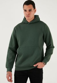 Zelená mikina s kapucňou a predným kengurím vreckom, vyrobená z jemného bavlneného zloženia, s rebrovanými manžetami a spodným okrajom, hladká textura.