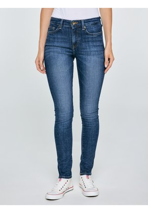 Niebieskie obcisłe dżinsy noszone z białymi trampkami, prezentowane od talii do stóp na jednolitym jasnym tle.