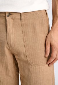 Ljusbruna byxor med fiskbensmönster, knappstängning och sidofickor. Tyget har en strukturerad yta och en avslappnad passform.