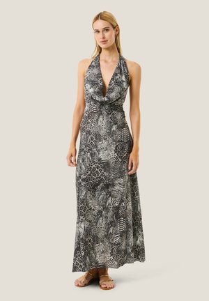 Donna con un abito senza maniche, collo halter, lungo fino al pavimento con stampa animalier grigia e nera, indossa sandali dorati, in piedi davanti a uno sfondo semplice.