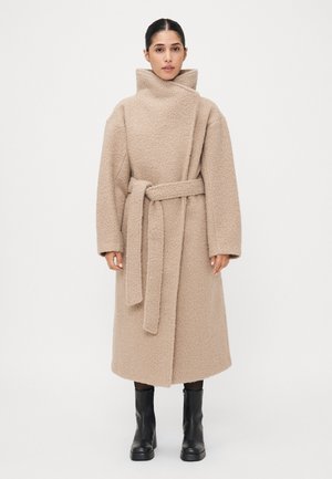 Beige kabát z vlněné směsi s vysokým límcem, zavinovacím designem a odpovídajícím páskem. Obsahuje texturovanou látku a spadlé ramena. Černé kotníkové boty.
