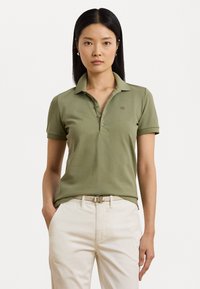 Polo vert olive à manches courtes, avec col et patte de boutonnage à trois boutons. Porté avec un pantalon crème et une ceinture claire sur laquelle figure un logo.