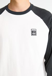 Bela majica z dolgimi rokavi z raglan rokavi z črnim vzorcem. Ima vezeno črno logo Adidas na prsih. Material je mešanica bombaža.