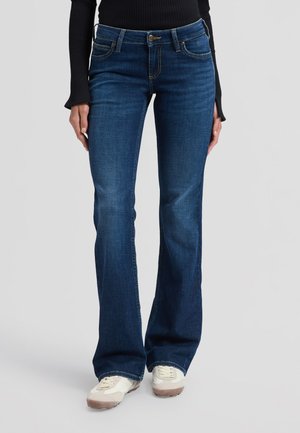 Femme portant un jean bootcut bleu foncé avec des baskets blanches et un haut noir à manches longues, debout devant un fond clair uni.