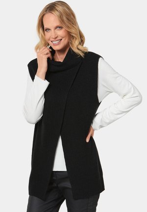 Cardigan sans manches en tricot noir avec un col en cowl, présentant un design ouvert à l'avant et une texture côtelée. Porté par-dessus un haut à manches longues blanc.