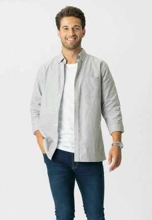 Nasmejani muškarac u svijetlosivoj košulji na kopčanje, bijeloj majici, plavim trapericama i ručnom satu, stoji s jednom rukom u džepu.