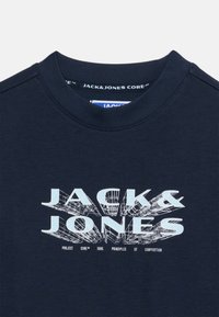 Μπλε ναυτικό μπλουζάκι με στρογγυλή λαιμόκοψη, με τη φράση "JACK & JONES" σε μεγάλα, ανοιχτό μπλε γράμματα 3D στυλ και μικρότερο κείμενο από κάτω στο εμπρός μέρος.