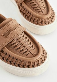 Brązowe, plecione buty typu slip-on z teksturowaną powierzchnią, wyściełanym paskiem i białą gumową podeszwą z wzorzystym brzegiem.