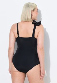 Costume da bagno intero nero con spalline regolabili, caratterizzato da un dettaglio arricciato su una spalla. Tessuto liscio e elastico con un design aderente.