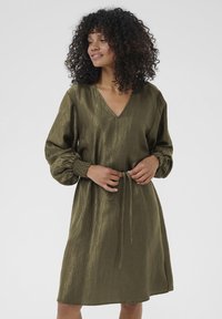 Femme aux cheveux bouclés portant une robe verte olive à manches longues avec un décolleté en V et une taille cintrée, souriant légèrement et regardant sur le côté.