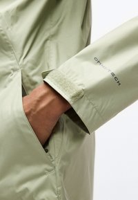 Χέρι στην τσέπη ενός ανοιχτό πράσινου αδιάβροχου μπουφάν με ρυθμιζόμενο μανσέτα βέλκρο, με ετικέτα "OMNI-TECH" στο μανίκι.