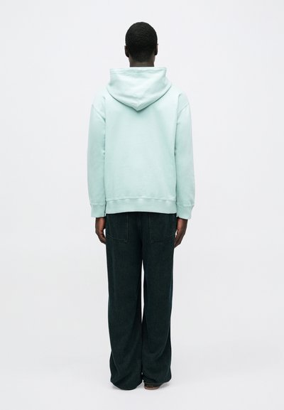 Helles mintgrünes Kapuzensweatshirt mit Kängurutasche, kombiniert mit weiten, dunklen Jeanshosen mit zwei Gesäßtaschen.