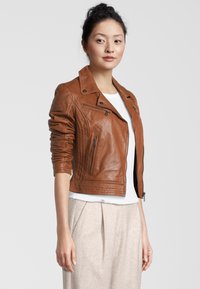 Bruine leren cropped bikerjack met gequilt details, ritssluitingen in de zakken en een opstaande kraag, gedragen over een wit shirt en lichte broek.