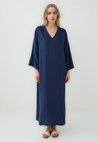Abito maxi blu navy con scollo a V, maniche larghe e una texture liscia, che arriva fino alle caviglie. Abbinato a sandali di colore chiaro.