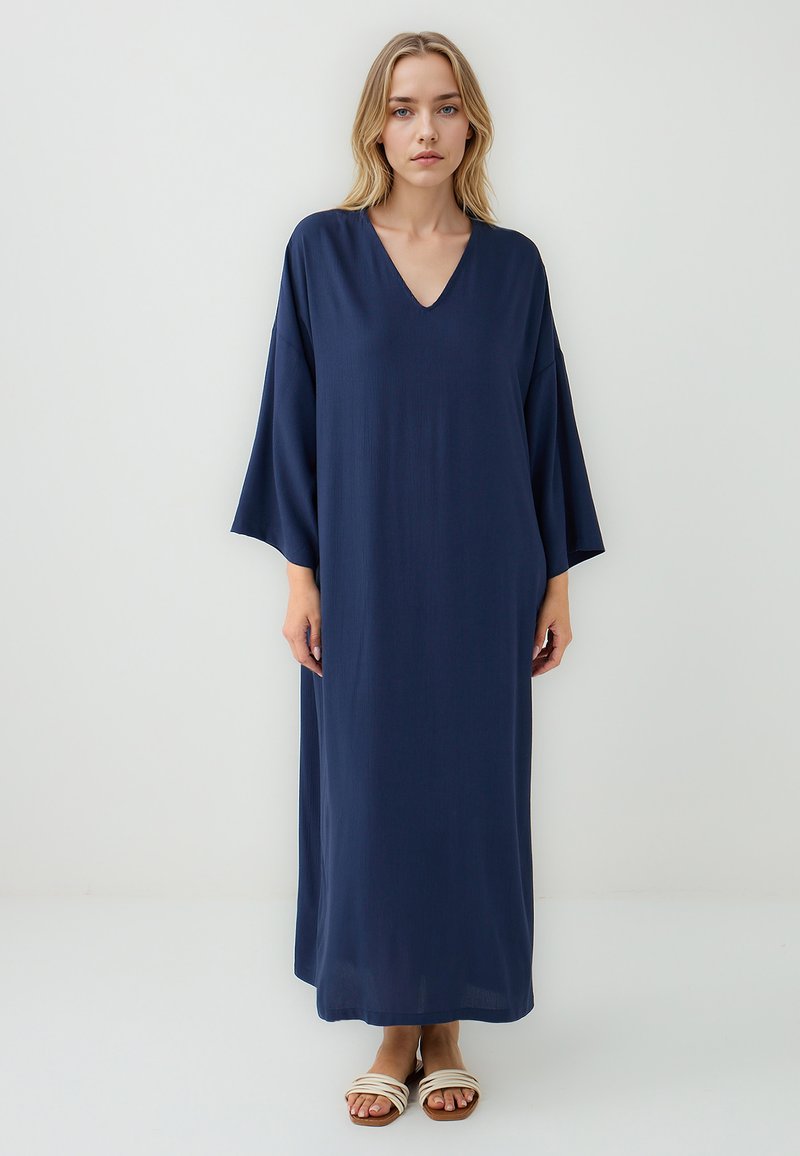 Navyblaues Maxikleid mit V-Ausschnitt, weiten Ärmeln und glatter Textur, das bis zu den Knöcheln reicht. Kombiniert mit hellen Sandalen.