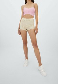 Beżowe szorty dżinsowe z postrzępionym dołem, zestawione z różowym krojonym topem. Modelka stoi w białych trampkach na neutralnym tle.