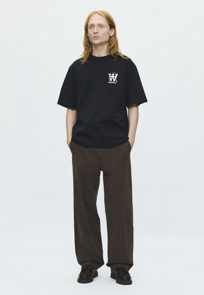 Jeune personne aux longs cheveux roux portant un t-shirt noir graphique, un pantalon marron ample et des chaussures noires, debout les mains dans les poches.