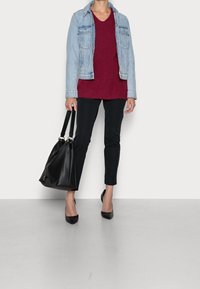 Jeansjacka över en vinröd stickad tröja, svarta byxor och svarta spetsiga klackar. En stor svart toteväska hålls i ena handen.