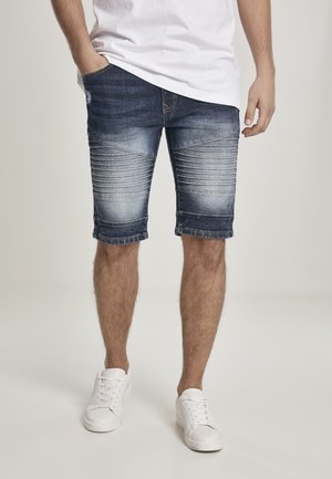 Shorts en denim bleu foncé avec un aspect usé, présentant des accents côtelés horizontaux sur les cuisses et des ourlets retroussés. Associés à des baskets blanches.