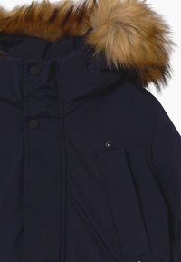 Marineblauwe parka met een capuchon afgezet met bont, drukknoopsluiting en een voorvak met een vlak ontwerp. Gladde textuur met gewatteerde isolatie.