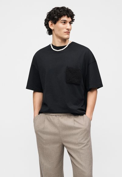 Νέος άντρας με κατσαρά μαλλιά φοράει μαύρο φαρδύ μπλουζάκι με υφή στην τσέπη, μπεζ παντελόνι και λευκό κολιέ με χάντρες, τα χέρια στις τσέπες.